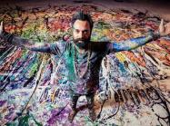 Artist Sacha Jafri covered in paint with arms spread, standing on a large paint-covered canvas
