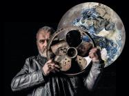 artist standing with metal fan in front of moon painting/ sculpture