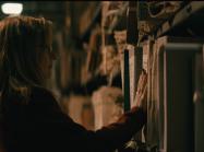Lynda is seen in dark room, looking at shelf packed with documents. 