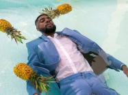 A man with dark skin, wearing a suit floats in a pool amongst three pineapples. 