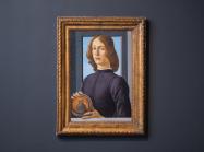 Botticelli's painting of a young man looking directly at the viewer hangs on a grey wall, in a gold frame. 