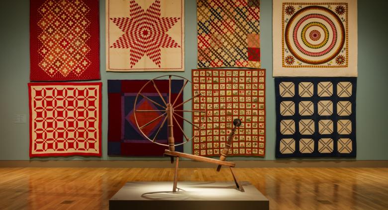 a gallery with eight quilts on the wall and a spinning wheel