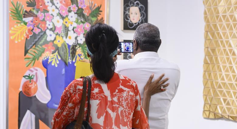 A couple at the Affordable Art Fair NYC, taking a photo of a bright, contemporary still life floral painting. 