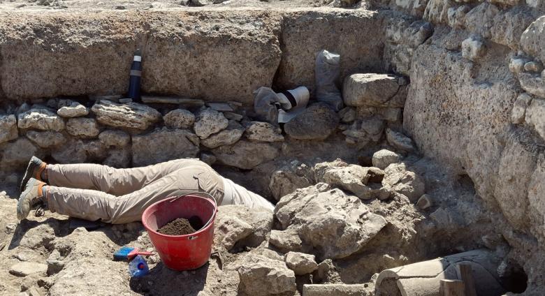 Excavation of certain physical features often requires archaeologists to get creative with digging methodologies. Wells, cisterns, and other subterranean features can be particularly challenging. Archeologist shown with head and torso in a hole as they work on site. 