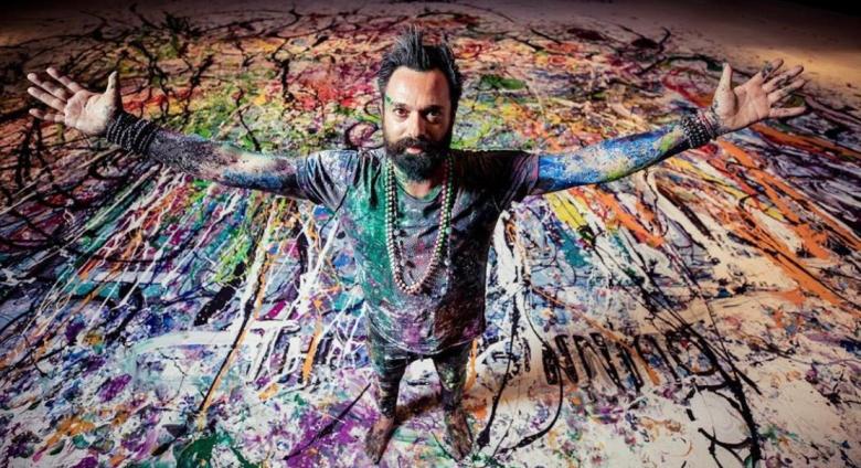 Artist Sacha Jafri covered in paint with arms spread, standing on a large paint-covered canvas