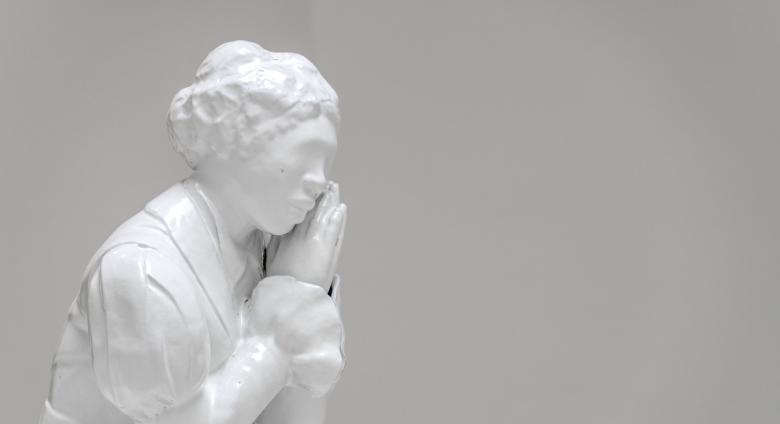 White material cast to show black female figure, dressed in clothes that seem to be from the 1800s, her hands are clasped together as if in prayer but held just to the side of her face to support her chin. 