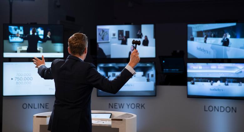 an auctioneer with a gavel stands in front of three large computer screens