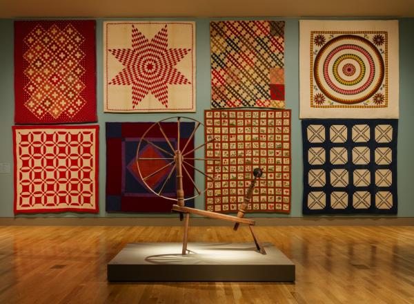 a gallery with eight quilts on the wall and a spinning wheel