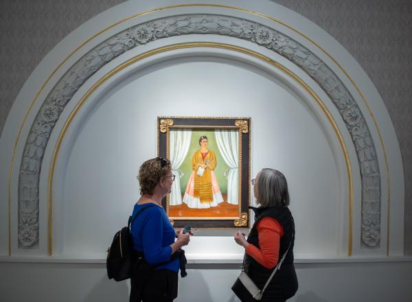 Two museum visitors are viewing a painting. The painting features a woman with a medium skin-tone wearing a dress and scarf and holding a piece of paper. It is framed by an arc with golden and floral ornamentation.