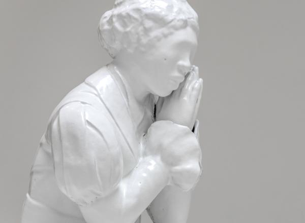 White material cast to show black female figure, dressed in clothes that seem to be from the 1800s, her hands are clasped together as if in prayer but held just to the side of her face to support her chin. 