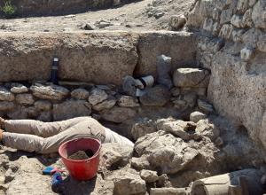 Excavation of certain physical features often requires archaeologists to get creative with digging methodologies. Wells, cisterns, and other subterranean features can be particularly challenging. Archeologist shown with head and torso in a hole as they work on site. 