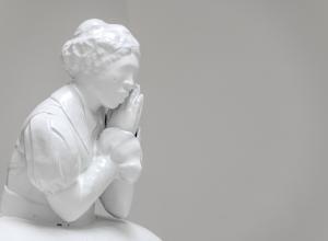 White material cast to show black female figure, dressed in clothes that seem to be from the 1800s, her hands are clasped together as if in prayer but held just to the side of her face to support her chin. 