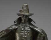 The figure looks directly at the viewer, with large hat and cape. The hat casts a shadow over part of the figure's face as detailed later. 
