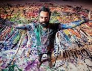 Artist Sacha Jafri covered in paint with arms spread, standing on a large paint-covered canvas