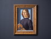 Botticelli's painting of a young man looking directly at the viewer hangs on a grey wall, in a gold frame. 
