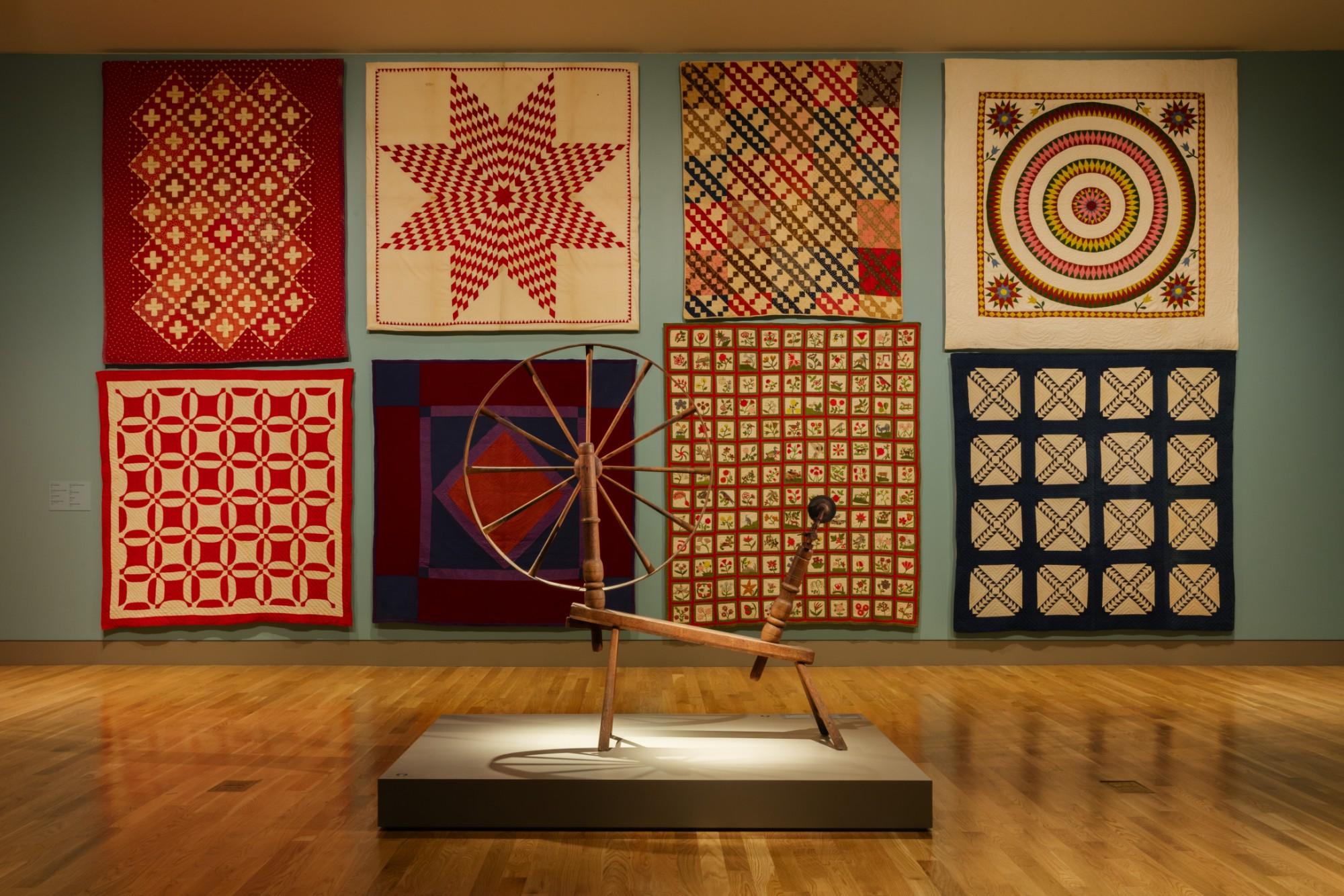 an art gallery with 8 quilts on the wall and a spinning wheel