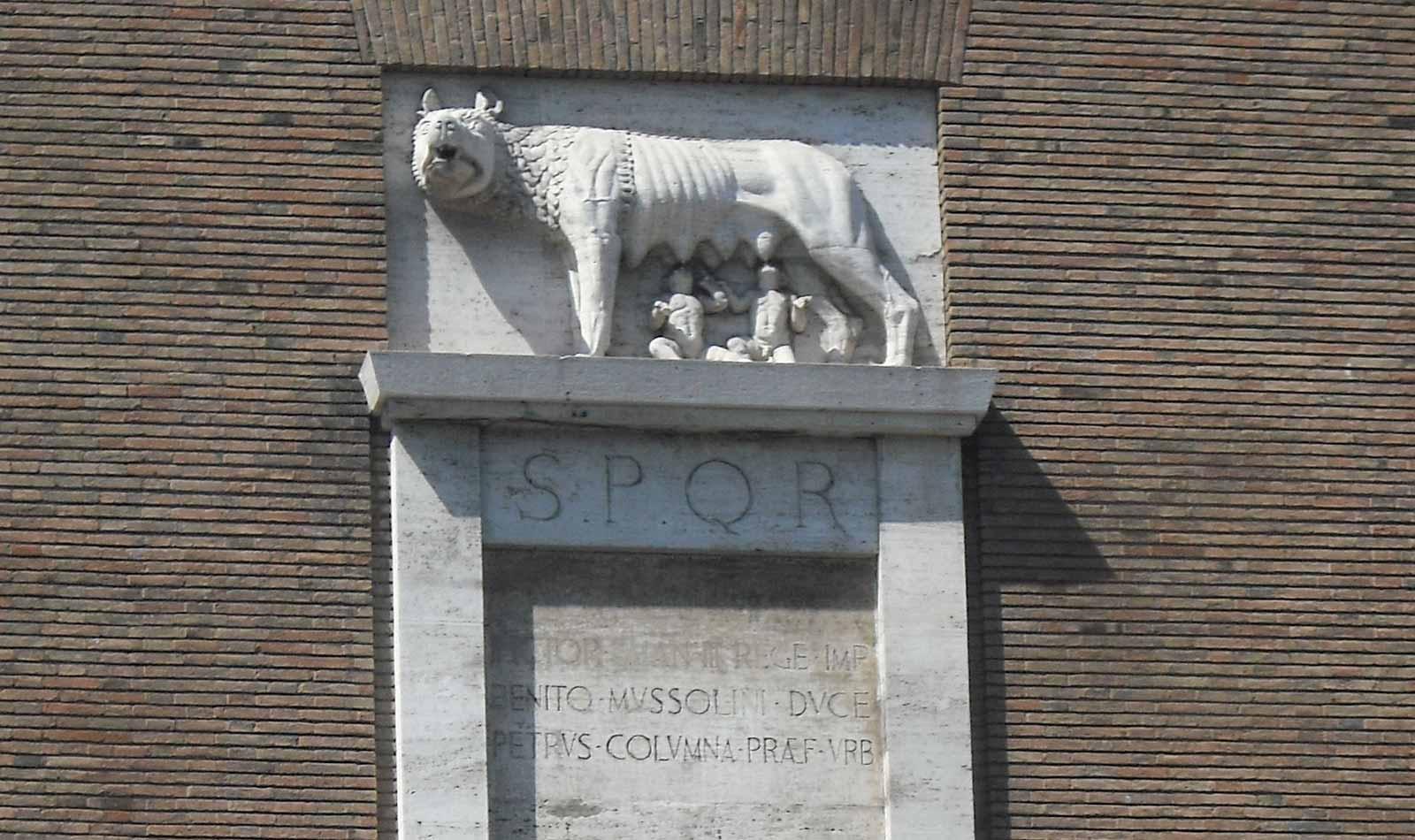Inscription with Mussolini's name still visible. The image of a She-wolf with twins, the symbol of ancient Rome, is above, but the three fasces below have been removed.