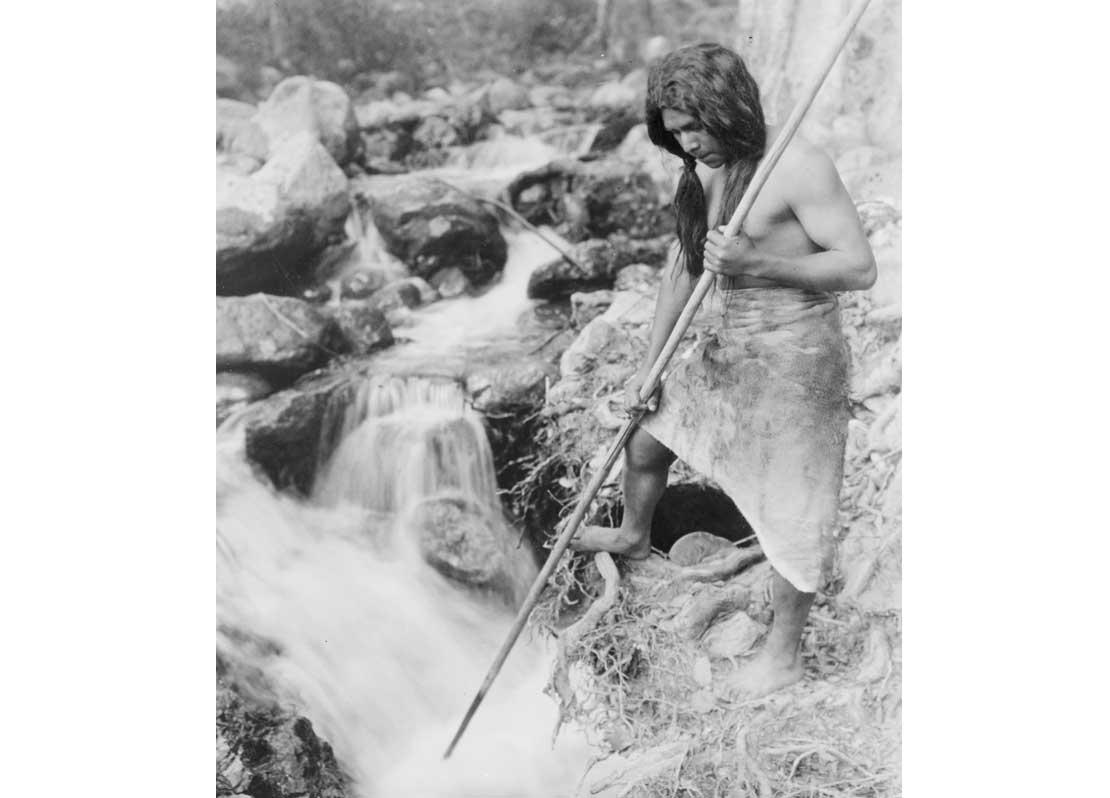 Hupa man with spear, standing on bank gazing into stream, 1923.