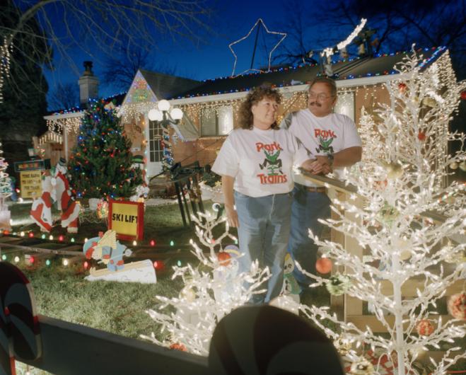 a couple in matching train t-shirts stand in front of their home decorated with christmas lights