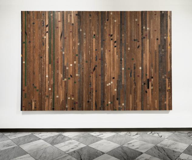 wall-mounted, flat artwork comprised of wood flooring arranged in a manner that emphasizes the color of certain planks of wood and creates an abstract, speckled piece. 
