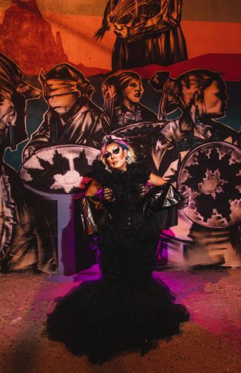 Lady Shug posed in an ally in a black dress, earrings that read land back and under red lighting. They stand in front of a mural of blindfolded native people. 