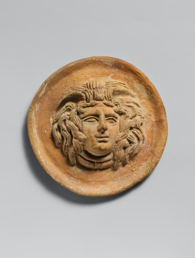 An aged circular terracotta slab depicting Medusa’s face and hair in the middle.
