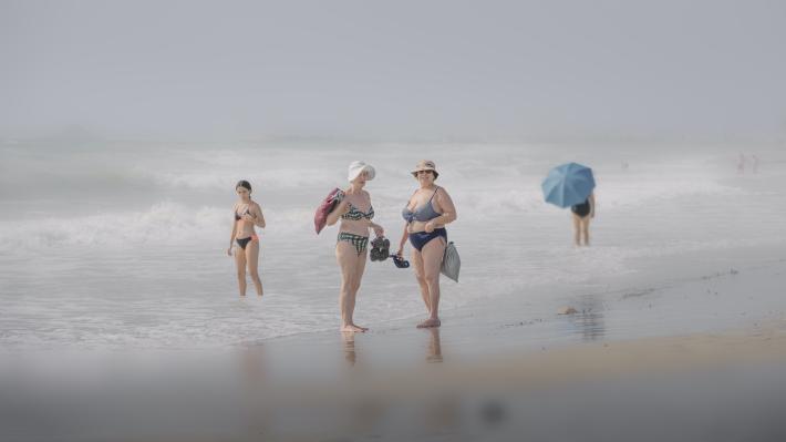 older women and younger woman on beach standing at edge of water in swim suits