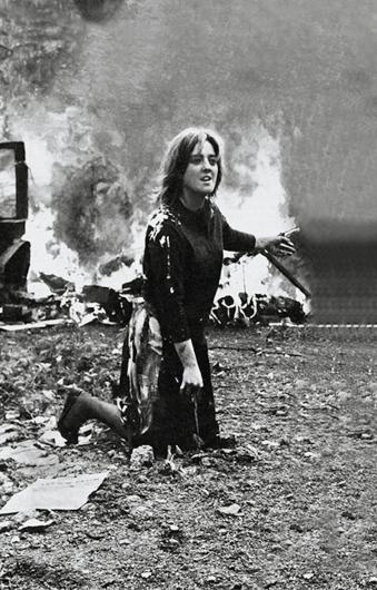 Marta Minujín, Photo from La destrucción (The Destruction), 1963. This photo is of a participatory art piece but it almost looks like a snapshot of a war scene. A woman kneels in the dirt, looking frantic while large objects burn behind her. 