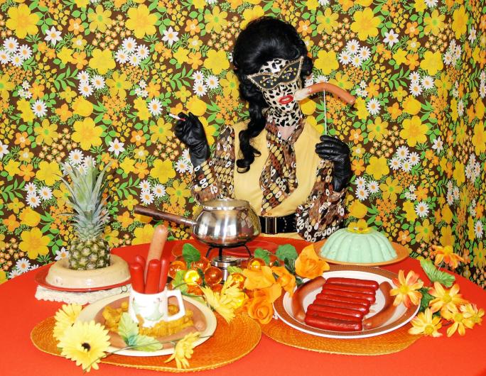 Miss meatface in neutral oranges and a leopard-print latex mask, sits in front of table loaded with pineapples, jello molds, and sausage plates. In front of busy floral background. 