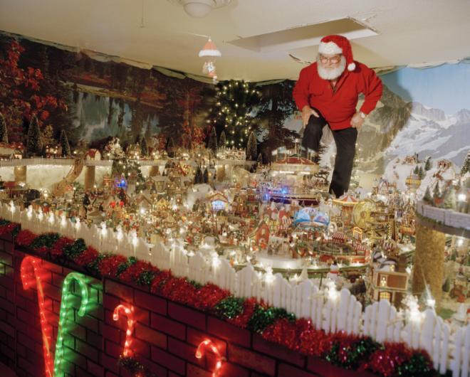 a man dresses as santa claus stands over a small model snowy village