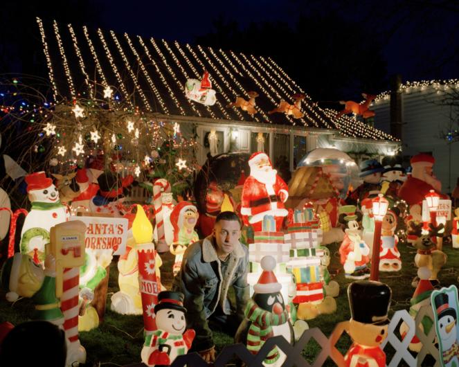 a young man crouches in front of a house with a yard full of inflatable and light-up christmas decorations