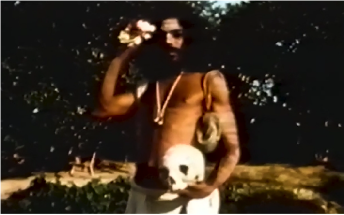film still showing a shirtless man with long hair holding a skull